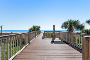 Ein Holzsteg führt zu einem Strand mit Palmen und blauem Himmel.