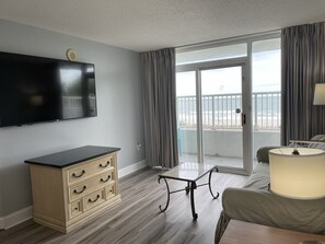 A hotel room with a flat-screen TV, a wooden dresser, a glass table, a sofa, and a balcony with a view of the beach.