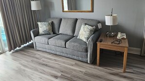 Un salon moderne avec un canapé gris, deux coussins assortis, une table d’appoint en bois avec une lampe, un téléphone et un miroir au mur.