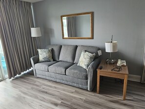 A modern living room with a grey sofa, two matching cushions, a wooden side table with a lamp, a telephone, and a mirror on the wall. - Oceanfront 3BR/2BA - Lazy River and Pools Resort (Myrtle Beach)