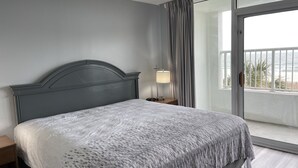 Une chambre à coucher comprenant un lit, une table de chevet, une lampe et une vue sur la mer, visible à travers une porte-fenêtre coulissante.