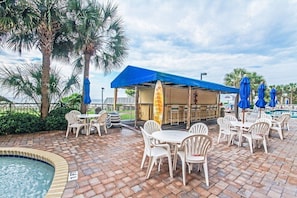An outdoor patio with white plastic chairs and tables, a pool, and a covered seating area with a blue roof. - Oceanfront 3BR/2BA - Lazy River and Pools Resort (Myrtle Beach)