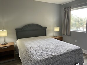 A bedroom with a bed, two bedside tables, a lamp on each table, a window with curtains, and a view of palm trees outside. - OCEANFRONT 3BR 2BA 1K1 Lazy River and Pools Resort (Myrtle Beach)