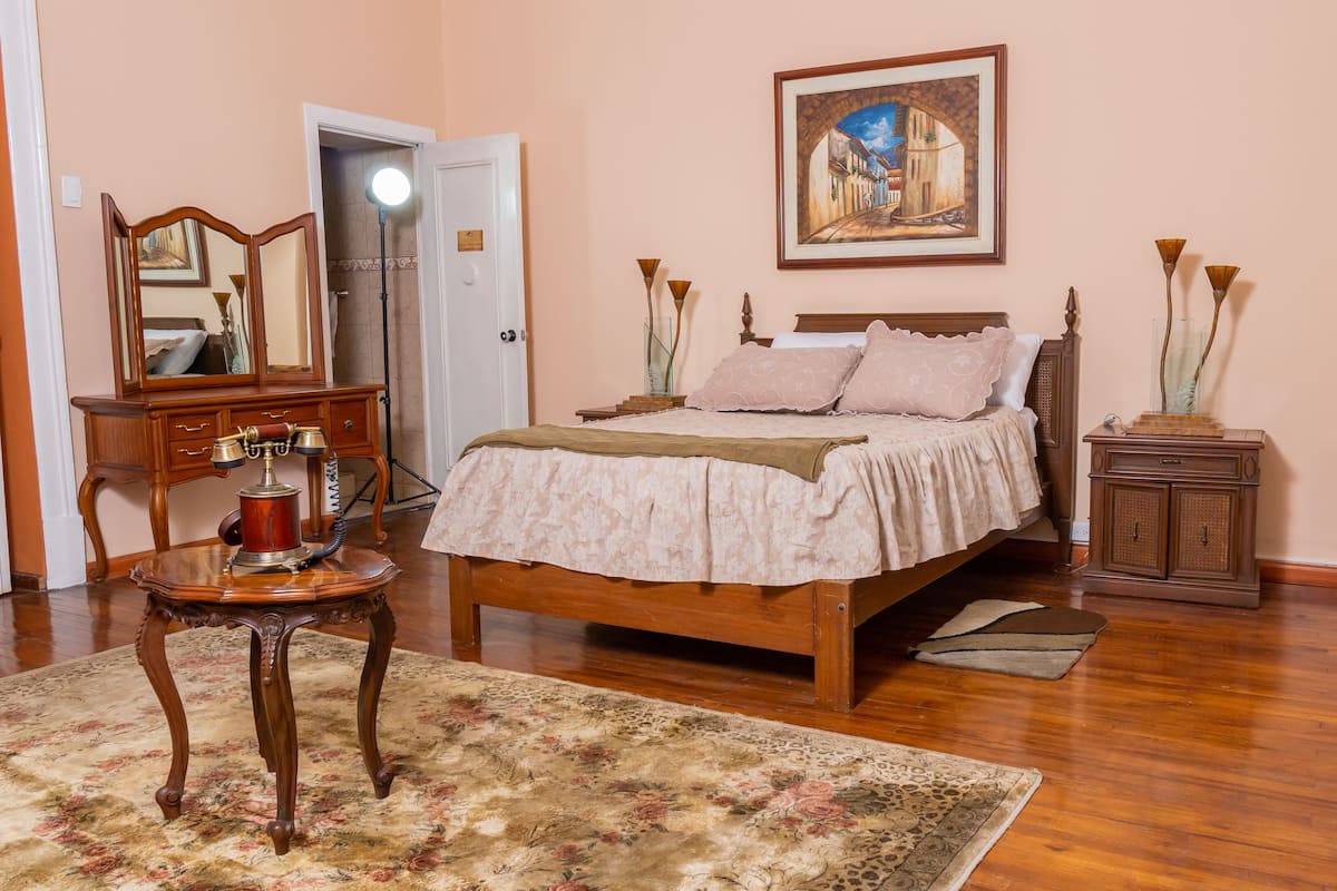 Une chambre à coucher avec un lit en bois, un bureau avec un téléphone, un miroir, un tableau accroché au mur et une petite armoire.