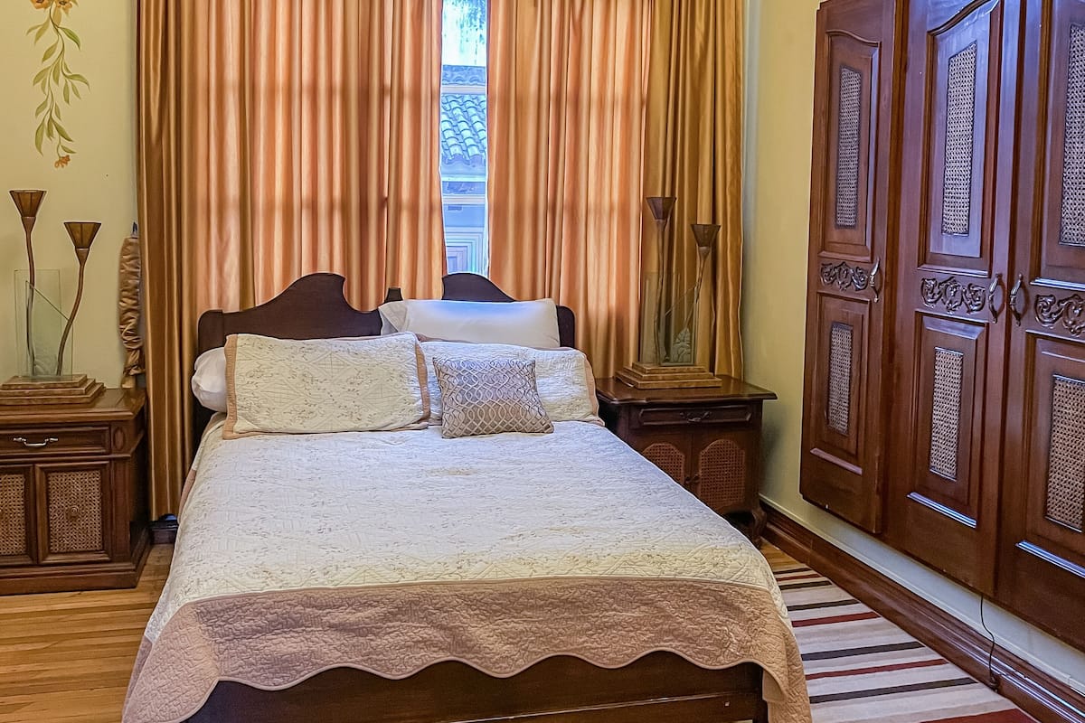 Une chambre avec un lit en bois, une grande fenêtre avec des rideaux, une armoire en bois et un mur décoratif.