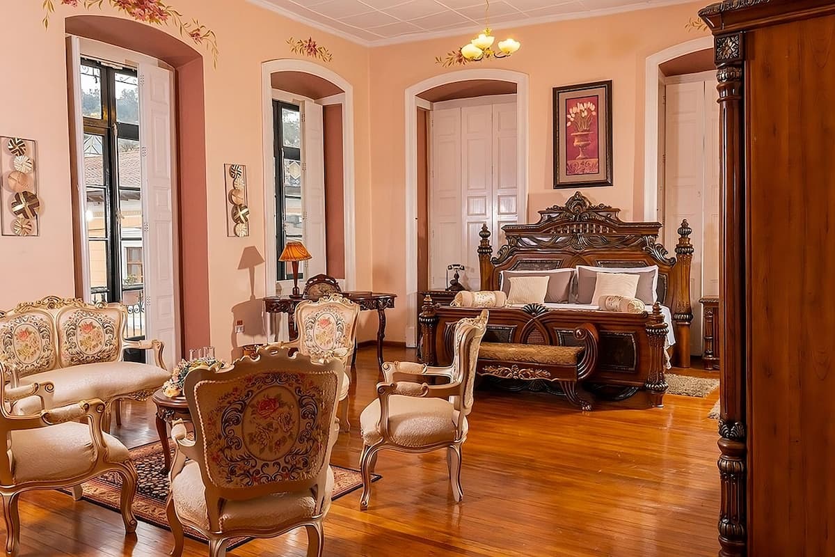 Une pièce meublée avec goût, comprenant un grand lit doté d’une tête de lit en bois, plusieurs fauteuils rembourrés et une table avec une lampe.