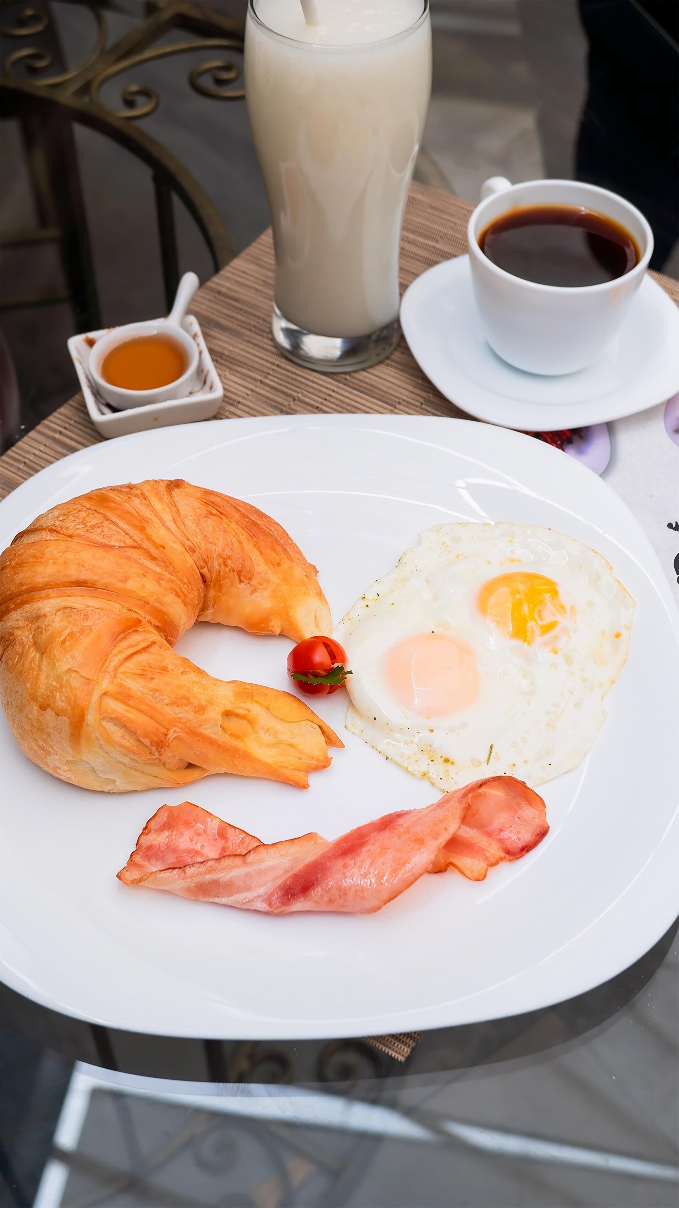 A breakfast plate with a croissant, fried egg, bacon, and a glass of milk.