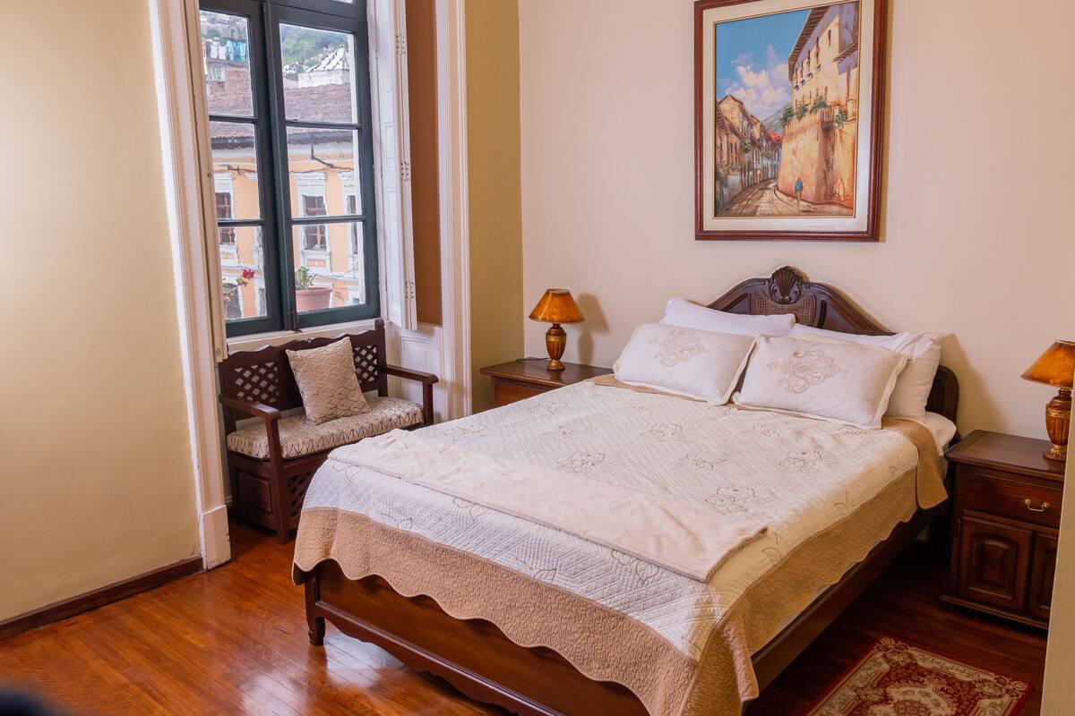 Une chambre à coucher bien aménagée, avec un grand lit, deux tables de chevet avec des lampes, un banc avec un coussin et une fenêtre donnant sur des immeubles.