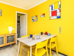 Une pièce lumineuse aux murs jaune vif, avec une table en bois et des chaises jaunes, une lampe blanche et une porte qui donne sur une autre pièce.