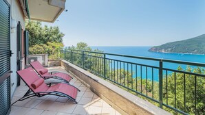 Ein Balkon mit zwei roten Liegestühlen, einem gefliesten Boden, einem Metallgeländer und Blick auf das Meer sowie eine entfernte Insel.