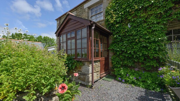 A traditional stone house with a wooden conservatory, surrounded by lush greenery and flowering plants.