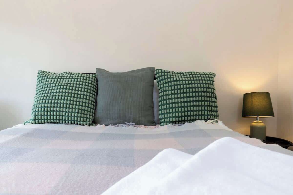 Un lit avec une couette blanche et des oreillers à motifs verts, une lampe sur la table de chevet et un mur uni.