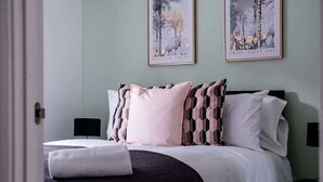 Una cama bien tendida con una colcha blanca y varios almohadones, una mesita de noche con una lámpara, y dos cuadros en la pared.