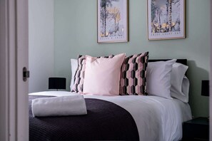 Un letto rifatto con un comofort bianco e diversi cuscini, un comodino con una lampada e due quadri appesi alla parete.