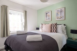 A neatly made bed with a dark patterned bedspread and pillows, a bedside table with a lamp, and two framed pictures on the wall. - Stylish 3 Bed Home With 2 Bathrooms & Parking (Bedford)