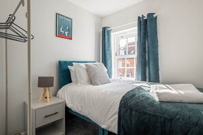A bedroom with a bed, a bedside table, a lamp, a window with curtains, and a poster on the wall.