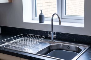 A kitchen with a stainless steel sink, a dish drying rack, and a window with a view of a brick wall. - Stylish 3 Bed Home With 2 Bathrooms & Parking (Bedford)