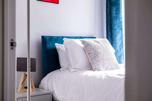 Une chambre à coucher avec un lit, du linge de lit blanc, une tête de lit bleue et une table de chevet avec une lampe.