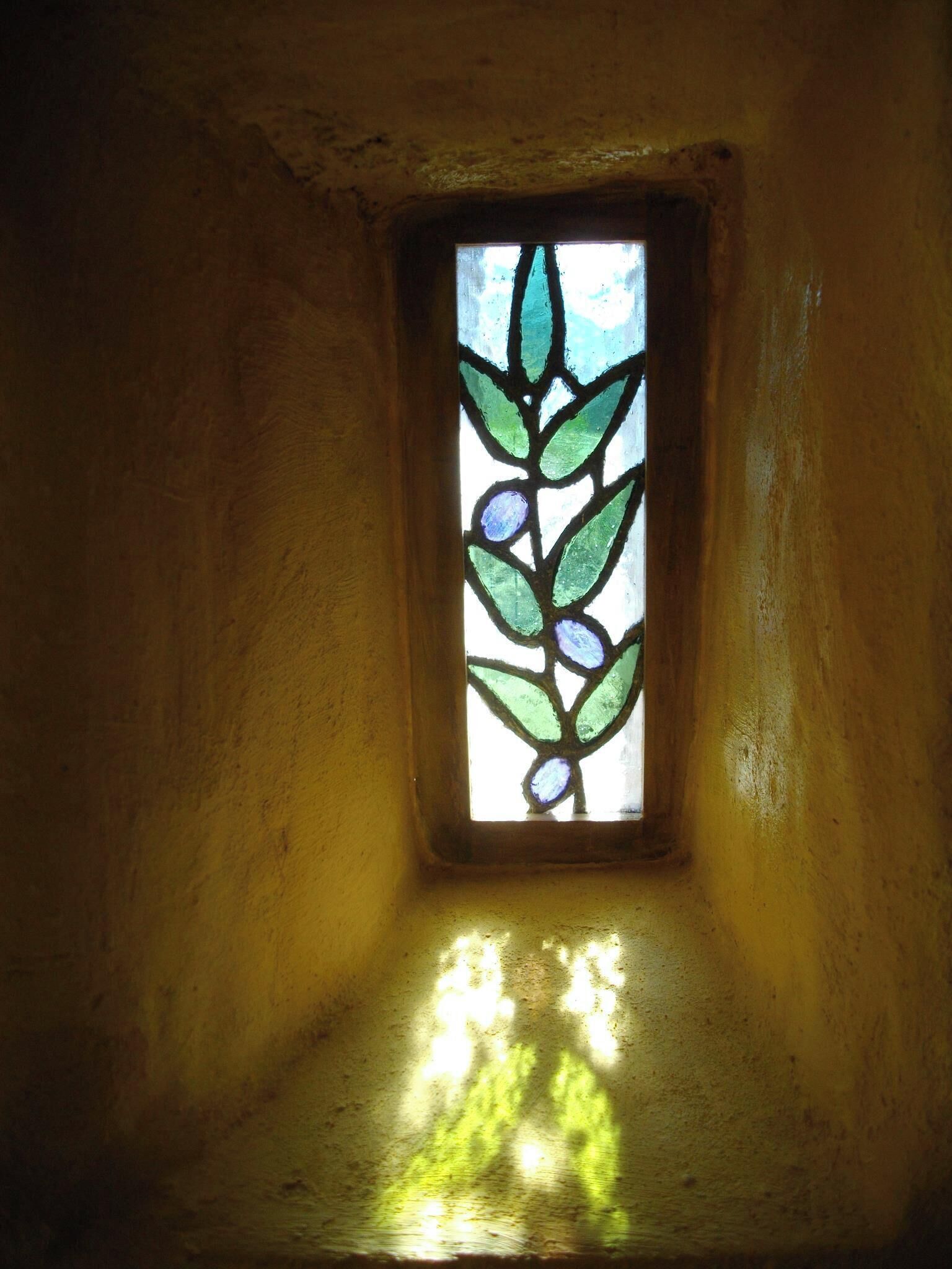 A small, square stained glass window with a leaf pattern, casting green shadows on the wall.