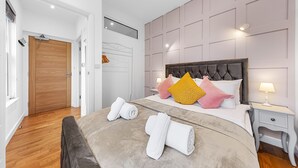 A hotel room with a wooden door, a bed with a tufted headboard, and a nightstand with a lamp. - Stunning 1 Bed Apartment Near Heathrow London (Staines)