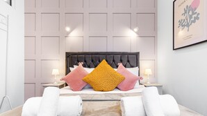 A hotel room with a tufted headboard, a beige carpet, and two bedside tables with lamps. - Stunning 1 Bed Apartment Near Heathrow London (Staines)