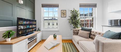 A modern living room with a flat-screen TV, a sofa, a coffee table, and a view of the city through large windows.