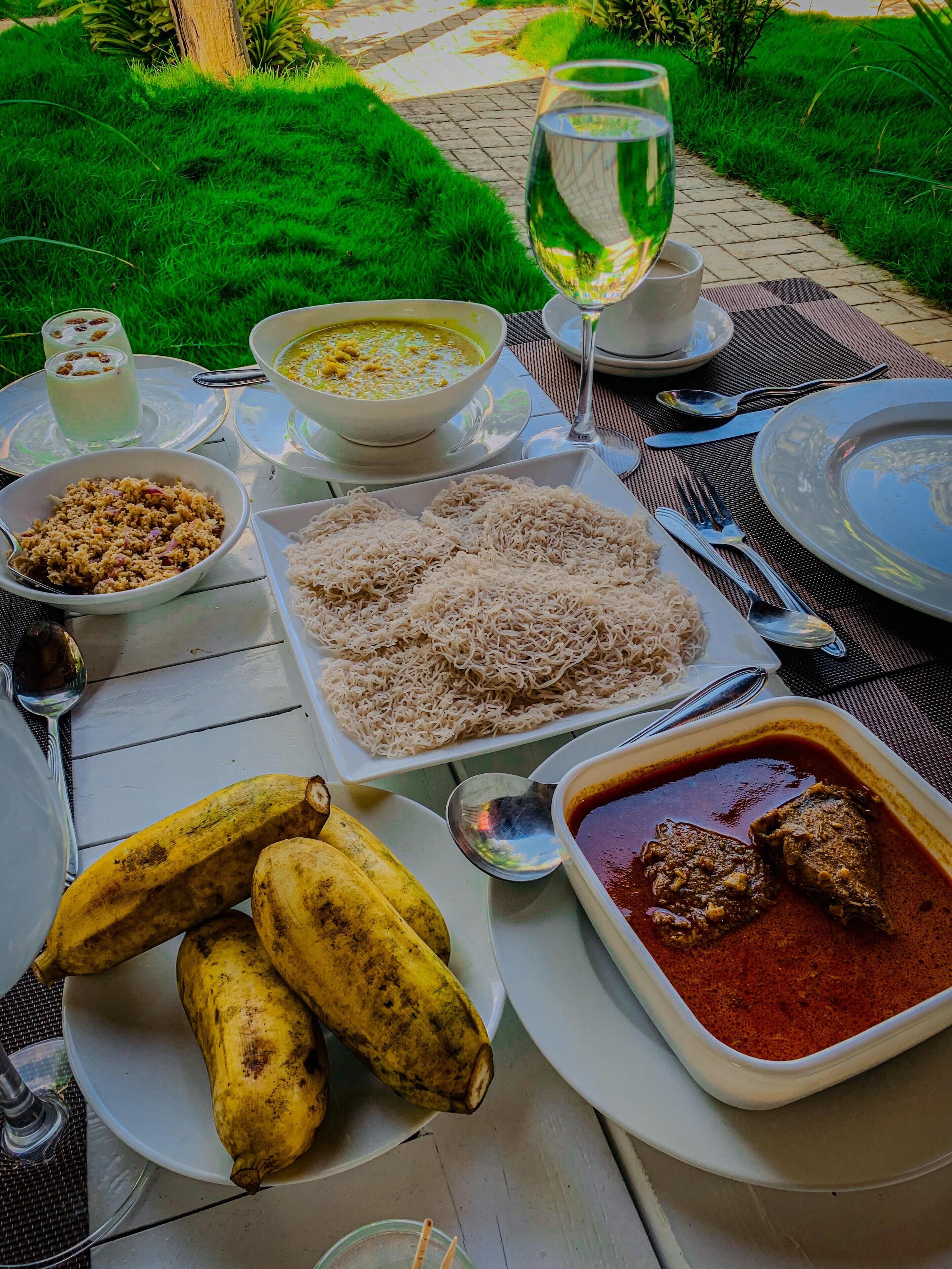 Une table dressée à l’extérieur, avec différents plats, notamment un bol de soupe, une assiette de nouilles et un plat contenant de la viande en sauce. On y trouve également des bananes et un verre d’eau.