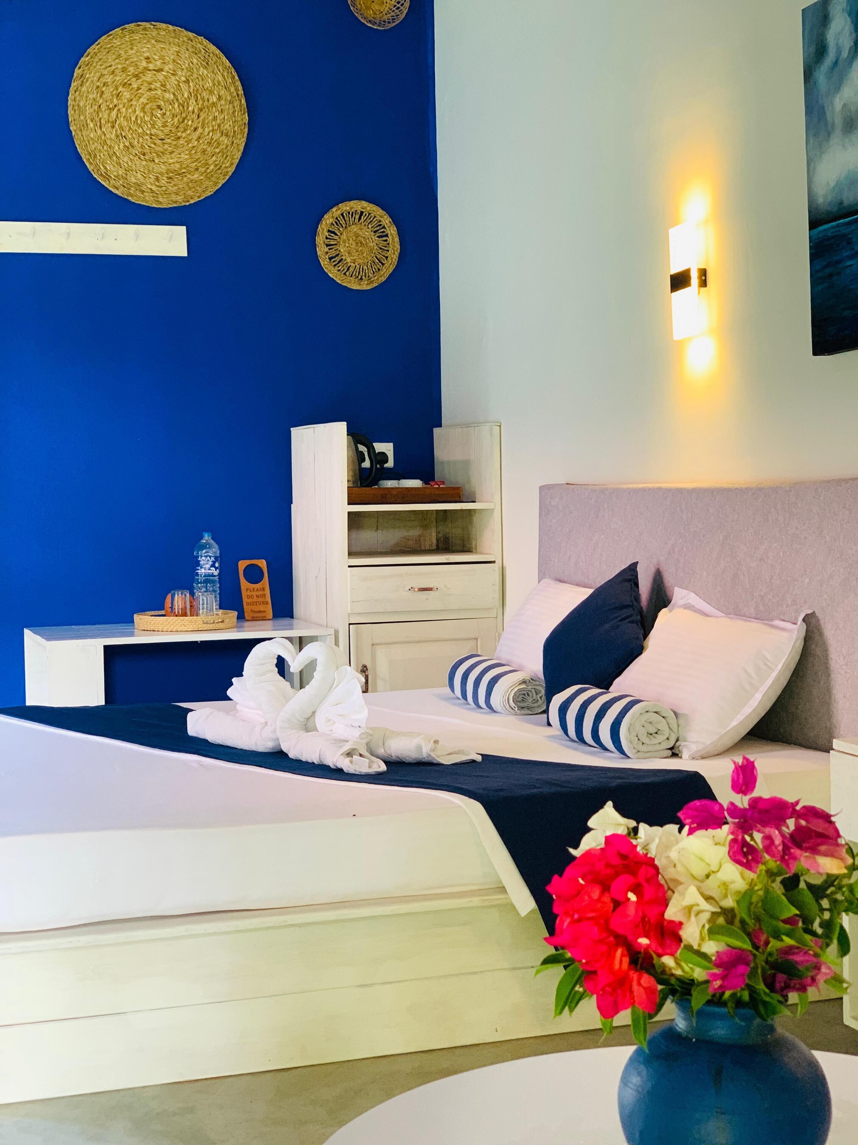 Een moderne hotelkamer met een blauwe accentmuur, een bed met wit en marineblauwe beddengoed, een nachtkastje met een fles en een mandje, een nachtlampje en een aan de muur bevestigd kunstwerk.