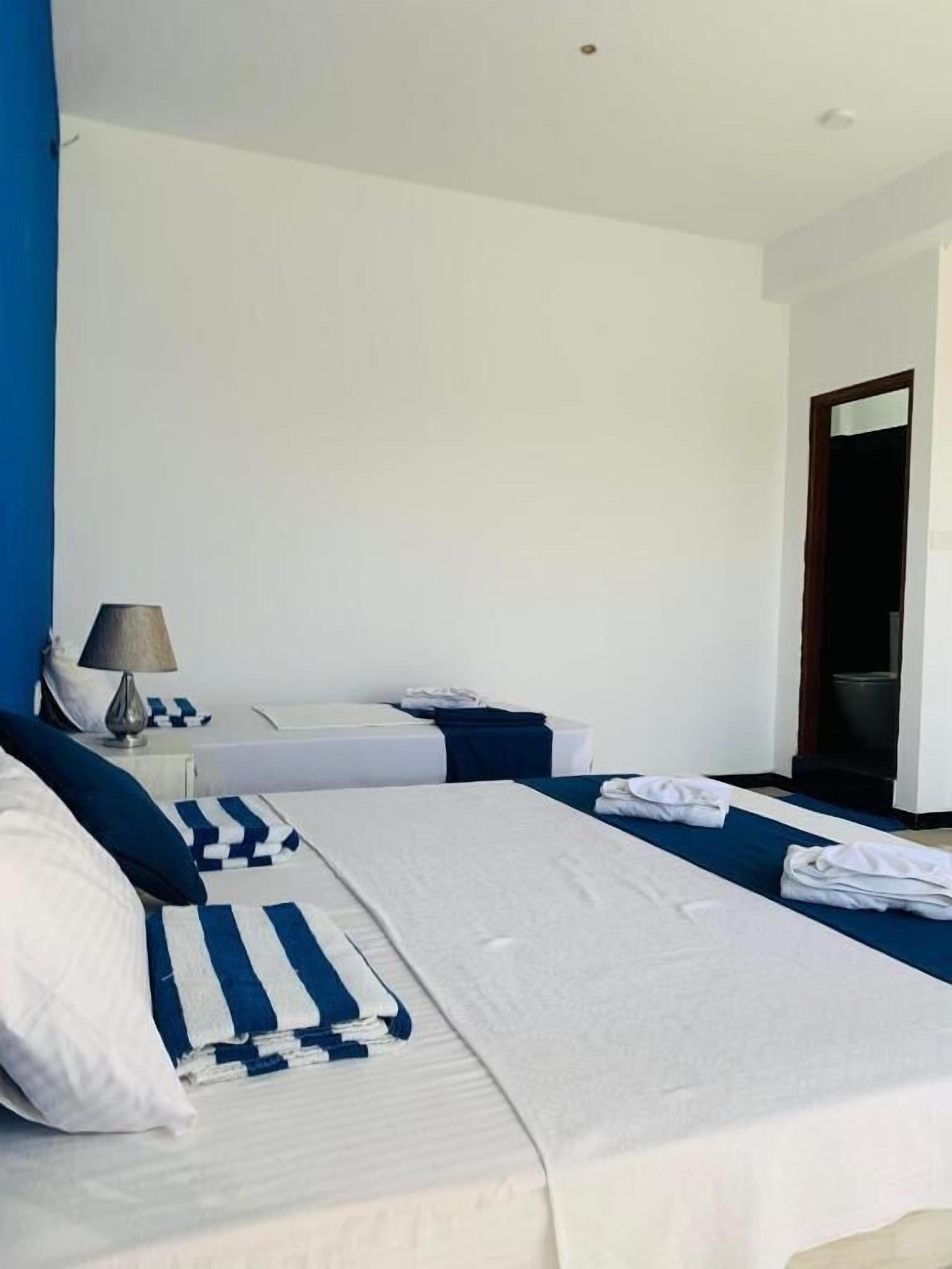 A hotel room with two beds, each with white bedding and blue accents, a nightstand with a lamp, and a door to the right.