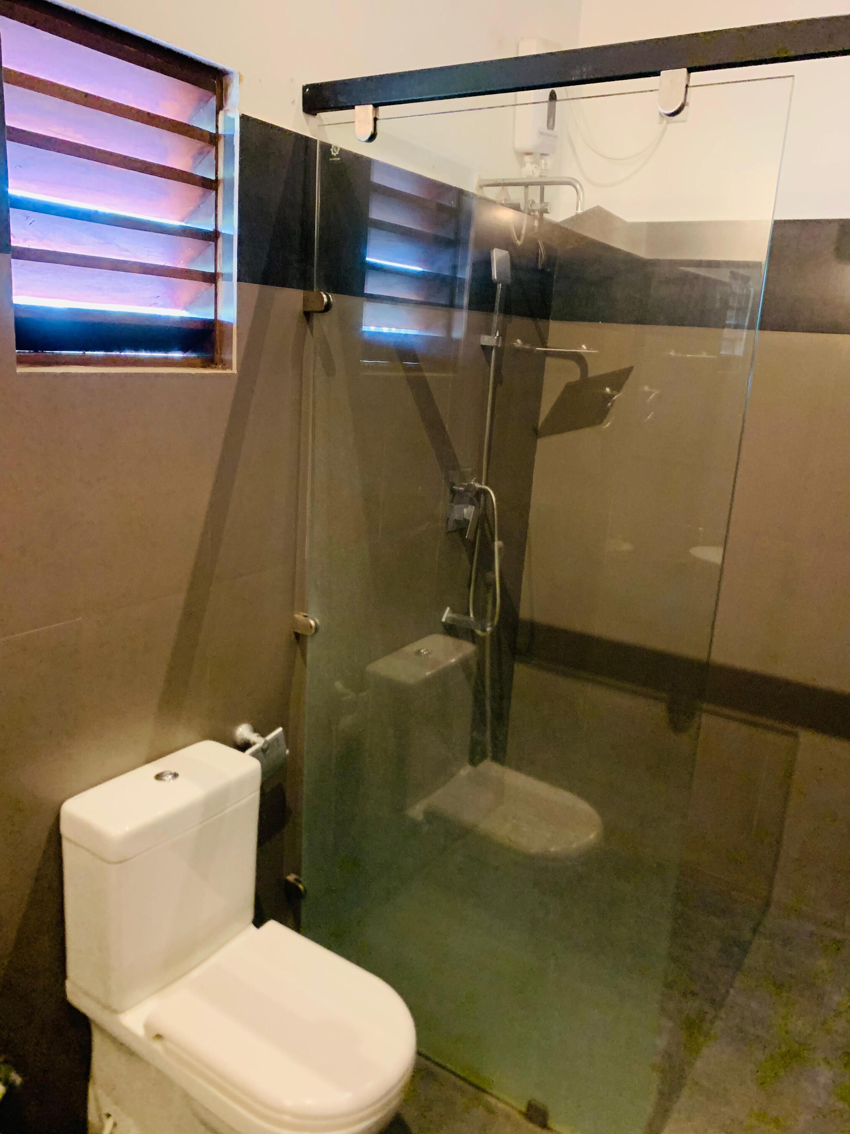 A bathroom with a glass-enclosed shower, a toilet, and a window with blinds.