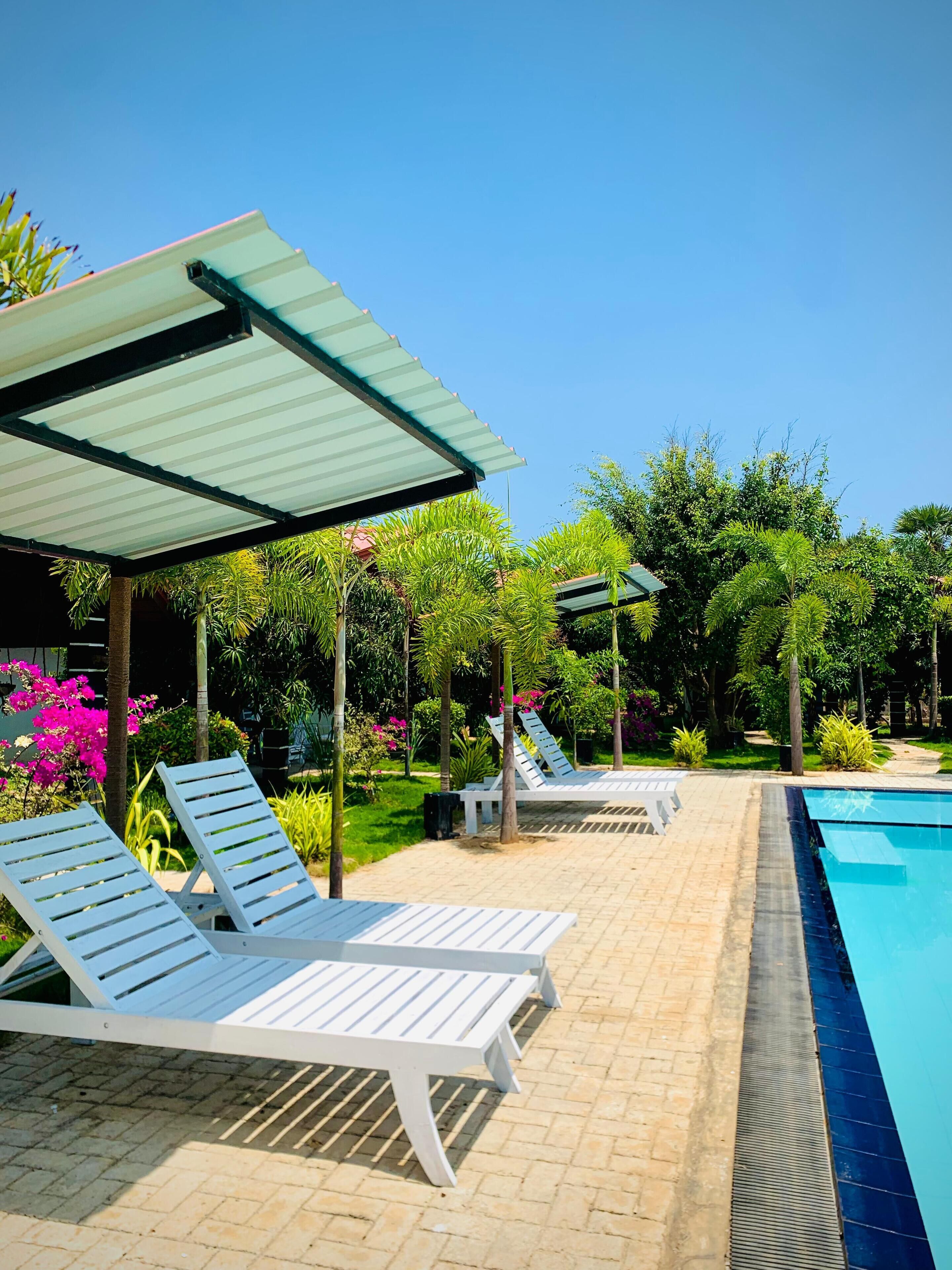 A poolside area with lounge chairs and a covered seating area, surrounded by lush greenery and a clear blue sky.