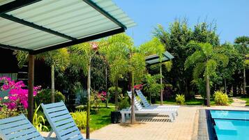 A poolside area with lounge chairs and a covered seating area, surrounded by lush greenery and a clear blue sky.