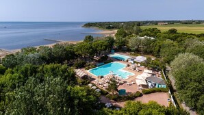 An aerial view of a resort with a large swimming pool surrounded by lounge chairs and umbrellas, a beach, and lush greenery.