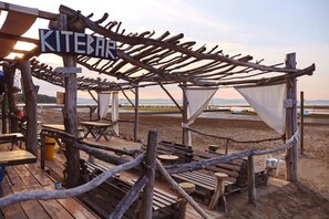 A rustic wooden deck with tables and chairs, a sign reading "KITEBAR," and a view of the beach and water in the background.