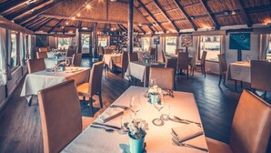 A restaurant with multiple tables set for dining, wooden flooring, and a thatched roof ceiling.
