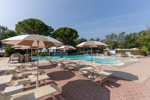 A pool area with lounge chairs and umbrellas, surrounded by trees and a clear sky.