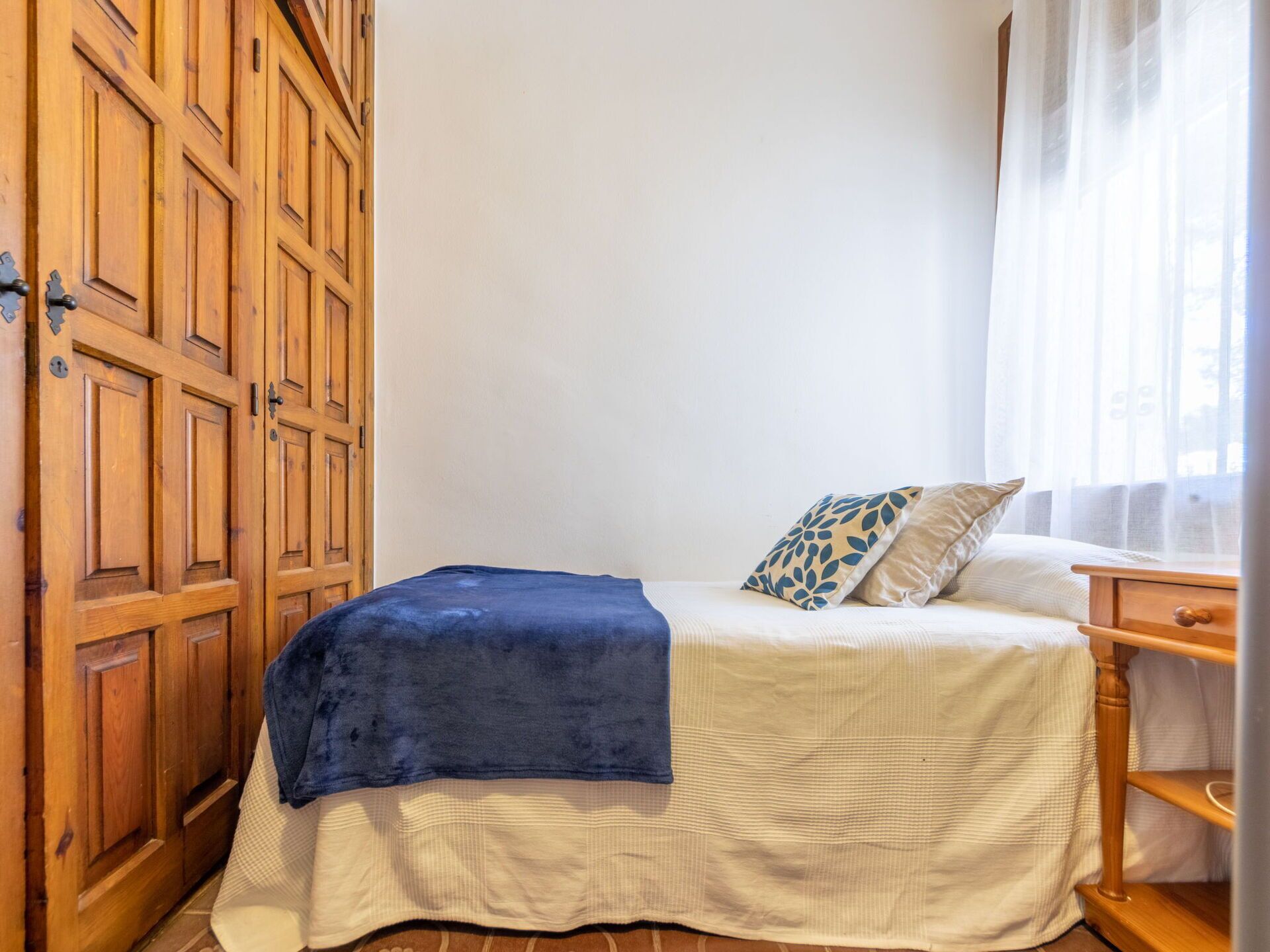 Une chambre à coucher comprenant un lit, une armoire en bois, une table de chevet et une fenêtre ornée de rideaux légers.