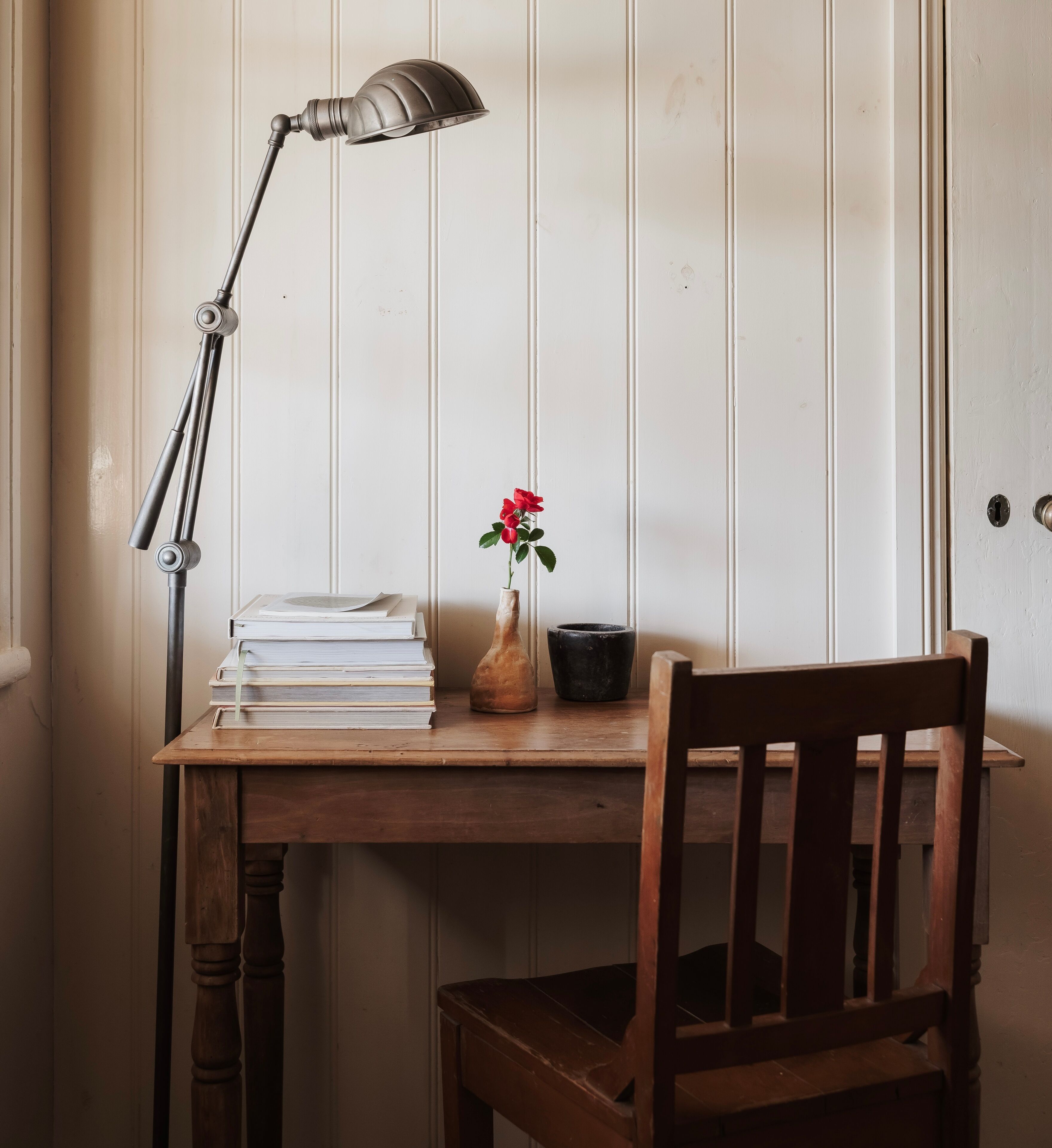 A wooden desk with a lamp, a vase with a flower, and a chair.