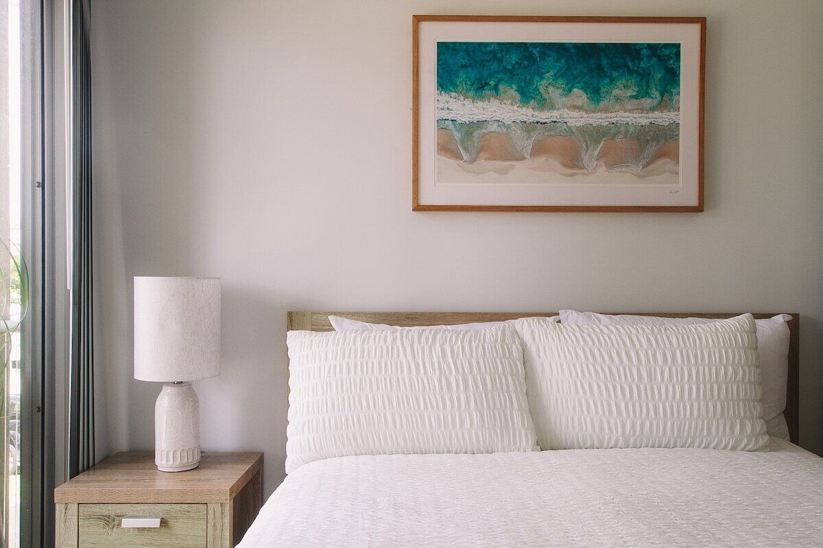 A bedroom with a bed, pillows, a nightstand with a lamp, and a framed painting on the wall.