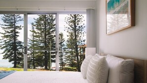 Une chambre avec un grand lit, une vue sur les arbres et un plan d’eau par une porte coulissante, et un tableau accroché au mur.