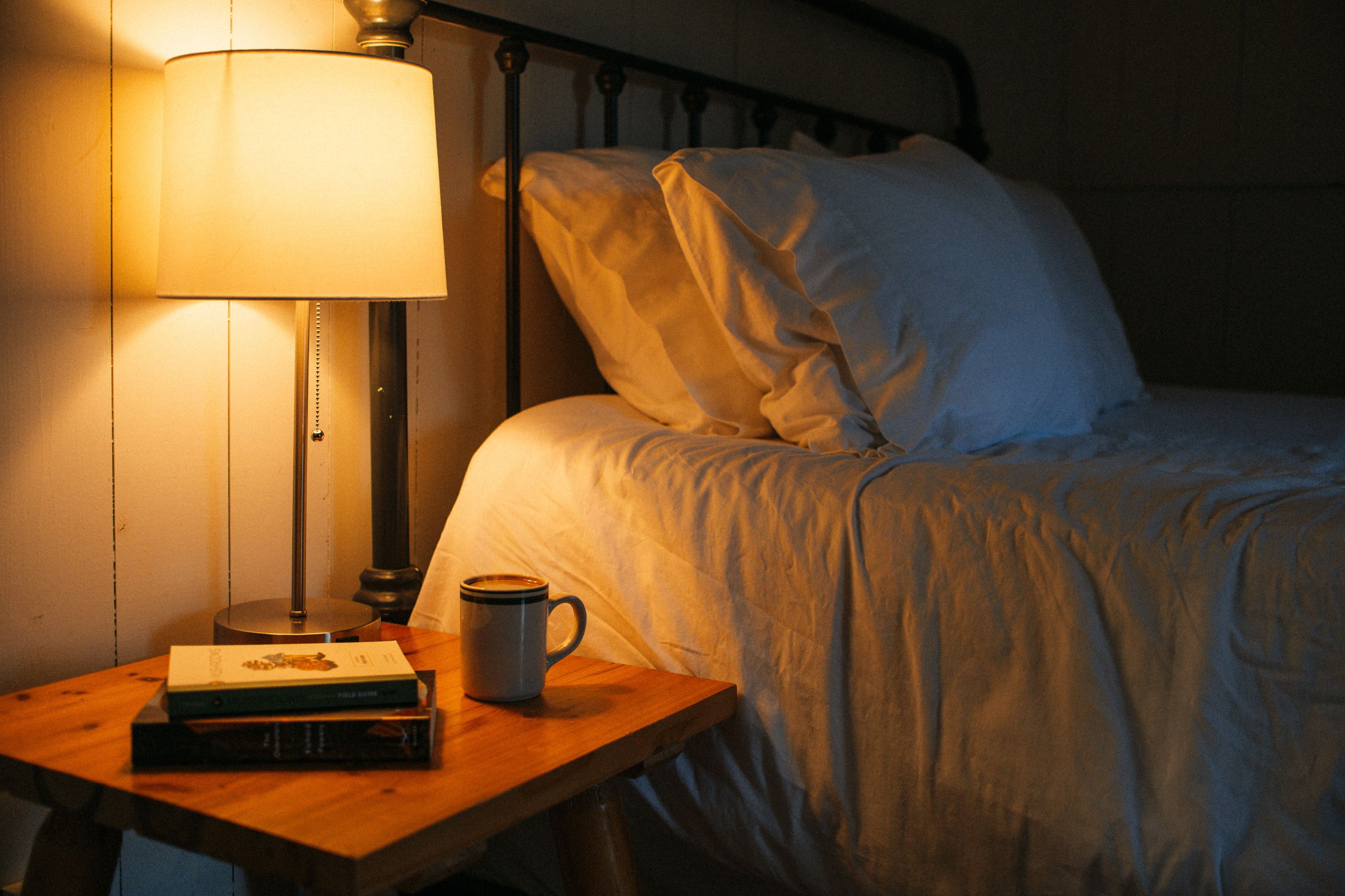 A neatly made bed with white linens, a bedside lamp, a cup, and a book on a wooden nightstand.
