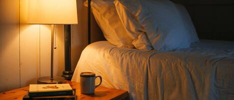 A neatly made bed with white linens, a bedside lamp, a cup, and a book on a wooden nightstand.