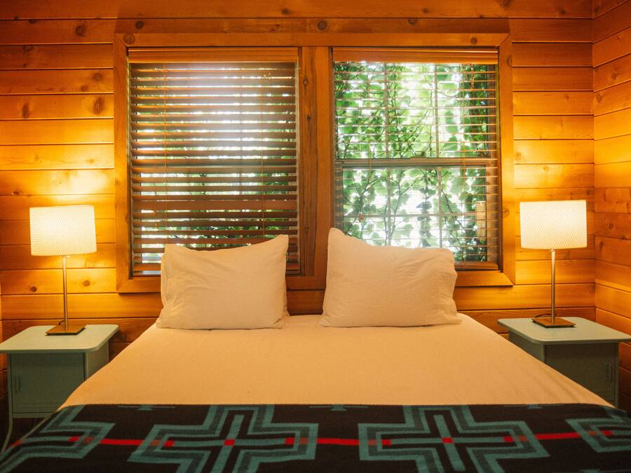 A wooden cabin room with a bed, two bedside lamps, and windows with blinds.