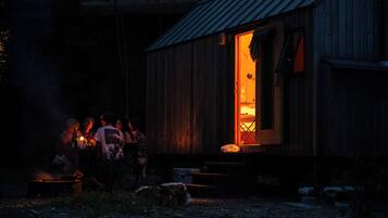 A rustic wooden building with a lit interior, a metal roof, and a small outdoor seating area with a fire.