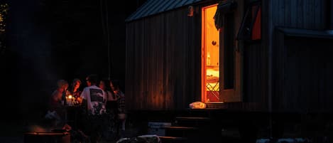 A rustic wooden building with a lit interior, a metal roof, and a small outdoor seating area with a fire.