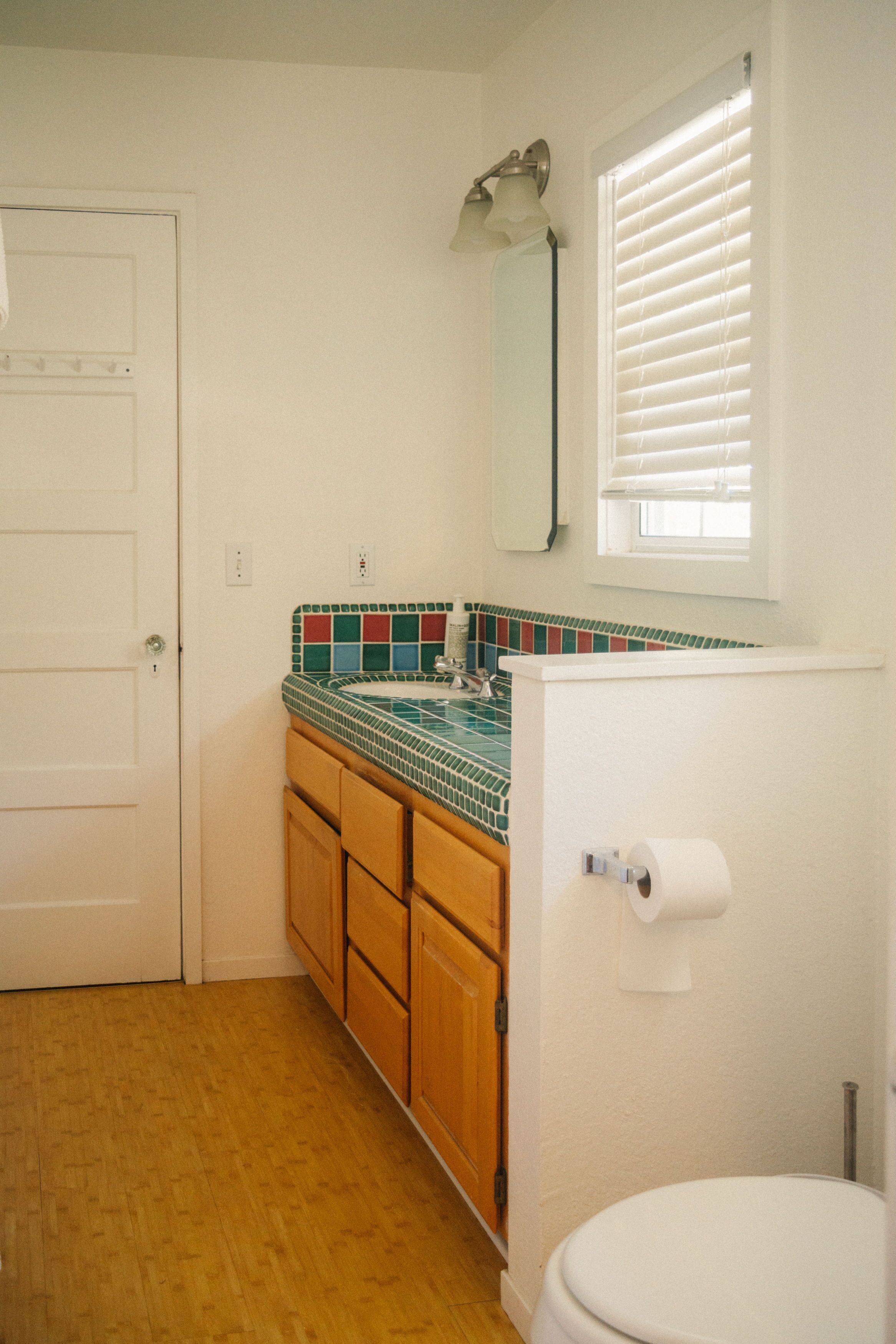 A bathroom with a wooden floor, a white toilet, a tiled sink with a green and red pattern, wooden cabinets, a white door, and a window with blinds.