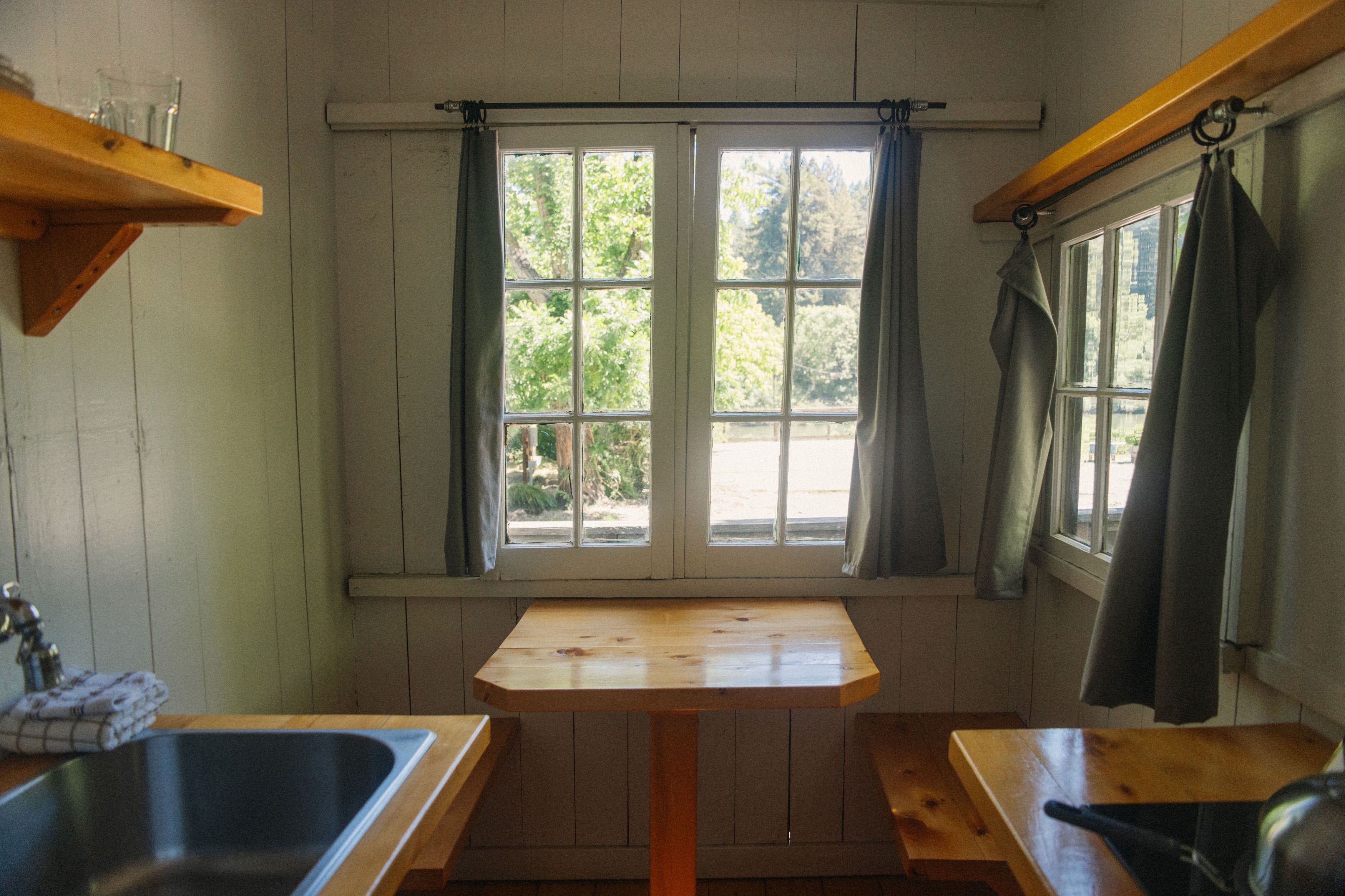 A small kitchen with a wooden table, two windows with curtains, a sink, and a wooden shelf above the sink.