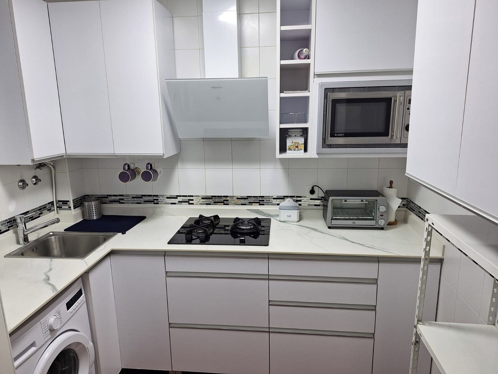 Une cuisine compacte avec des meubles blancs, un four à micro-ondes encastré, un grille-pain et un lave-linge.