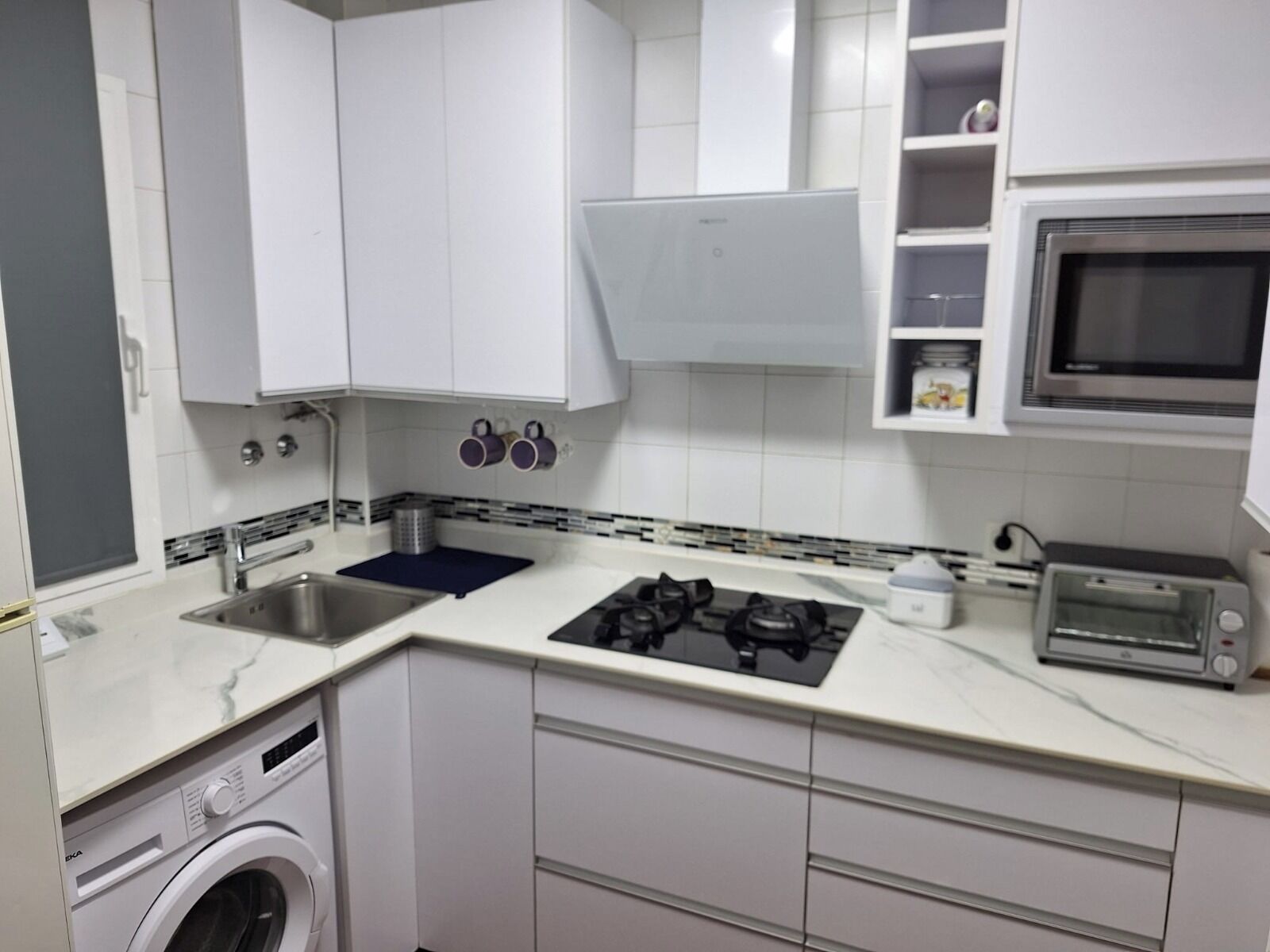 Een compacte keuken met witte kastjes, een ingebouwde wasmachine, een gasfornuis, een magnetron en een broodrooster.