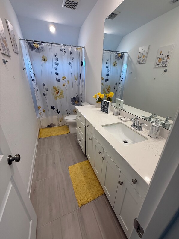 A modern bathroom with a white sink, mirror, and toilet. There is a yellow rug on the floor, and a shower curtain with a floral pattern. - Cozy & New 4Bd/ 3ba, in quiet neighborhood w/big yard. (Houston)